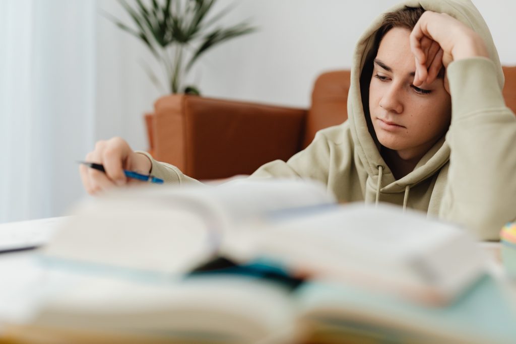 Adolescente estudiando con libros abiertos y una mano apoyada en la frente.